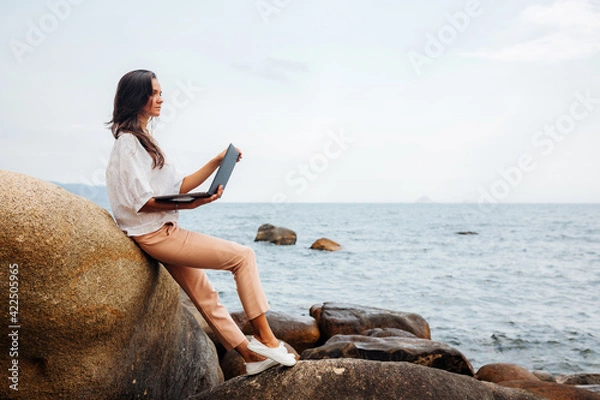 Obraz Young successful girl working using a laptop, sitting on a rocky seashore. traveling, blogger, freelancer, content plan, work online