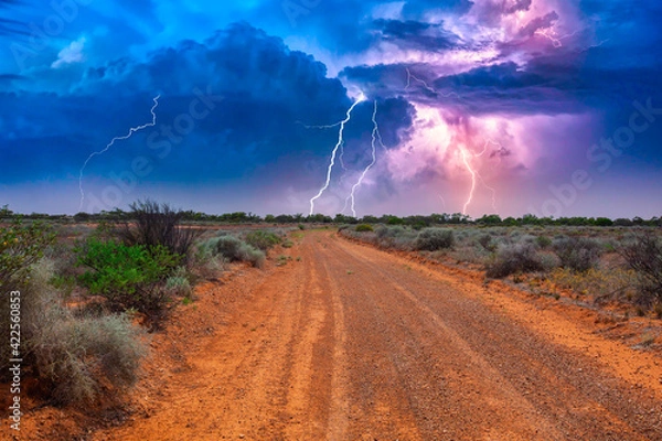 Fototapeta Deserted Australian outback landscape with red dirt road towards horizon with bushes in roadsides and heavy thunderstorm with white purple lightnings on the horizon