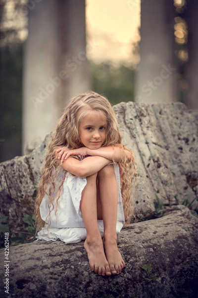 Fototapeta Portrait of beautiful blonde long-haired girl sitting on the ruined stones during the sunset in Pavlovsk park, Russia. Image with selective focus and toning