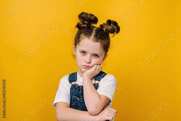 Obraz Serious pensive little kid girl looking unhappy at camera, tired upset preschool child with thoughtful face holding hand under chin, melancholic or bored, isolated over yellow studio background