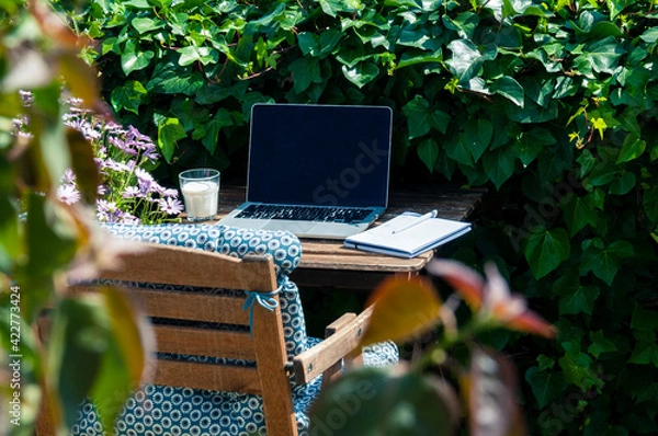 Fototapeta Remote workplace, laptop, notebook with a pen, a glass of milk are on a wooden table, next to a chair with a blue pillow, all this is in the green garden with blurred leaves, not the foreground