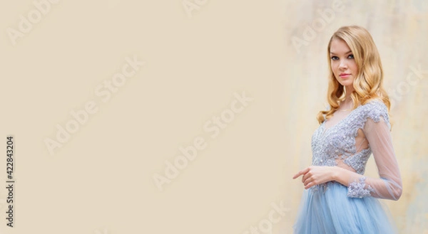 Obraz Portrait of a young blonde woman in a delicate blue wedding dress against a beige wall