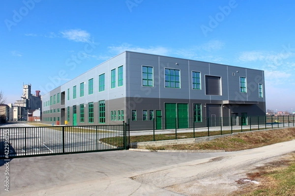 Fototapeta Newly built modern grey and green production industrial building surrounded with closed metal fence and large old concrete storage silo building in background on warm sunny winter day