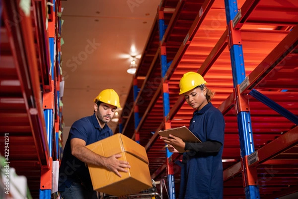 Fototapeta warehouse workers logistic team wearing hardhats to working in aisle between tall racks with packed goods, While inspecting industrial plants and warehouses for international shipping businesses