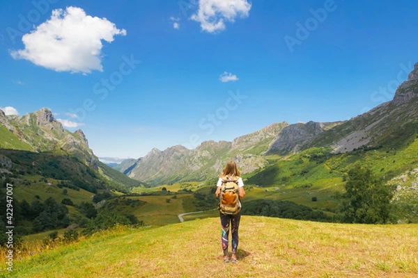 Fototapeta A girl with a backpack is looking to the mountains and there is a blue sky and some white clouds