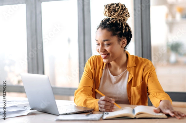 Obraz Edukacja online, e-learning. Szczęśliwa afroamerykańska młoda kobieta w stylowych swobodnych ubraniach, studiując zdalnie, używając laptopa, słuchanie wykładu online, robienie notatek podczas siedzenia w domu, uśmiecha się
