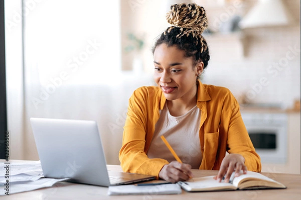 Obraz Skoncentrowana urocza stylowa afroamerykańska studentka z afro dreadlocks, studiując zdalnie z domu, używając laptopa, notatki na temat notatnika podczas lekcji online, koncepcji e-learningu, uśmiechnięcia się