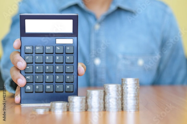 Obraz A businessman with a calculator and a stack of coins in foreground. Business, finance and growth concept