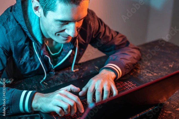Fototapeta A male hacker is typing on a laptop keyboard in a dark room under a neon light. Cybercrime fraud and identity theft