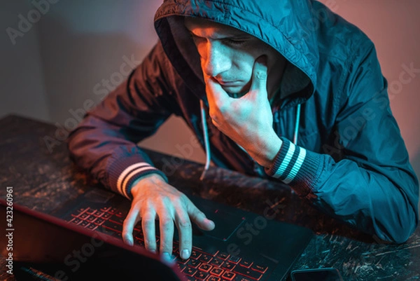Fototapeta Hooded hacker is typing on a laptop keyboard in a dark room under a neon light. Cybercrime fraud and identity theft