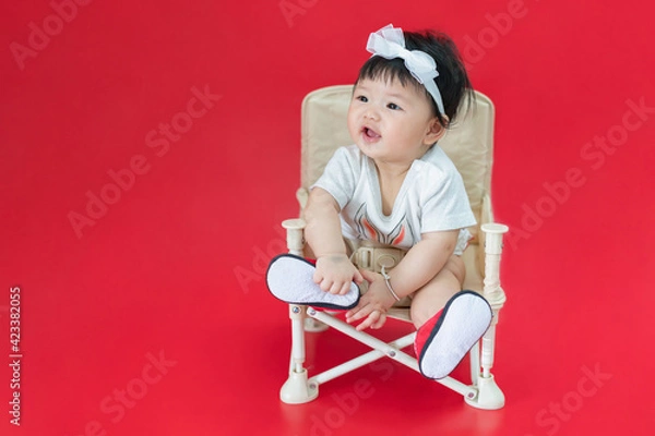 Fototapeta Asian happy baby smiling and sitting on a chair in red color background. Cute 6 months baby with copy space use as concept of bedroom, drypers, development, baby or kid department in hospital.