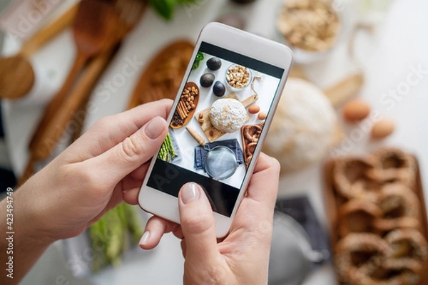 Fototapeta Food blogger taking a picture with her phone of groceries on white table. Picture of ingredients for breakfast on smartphone. Blogging concept.