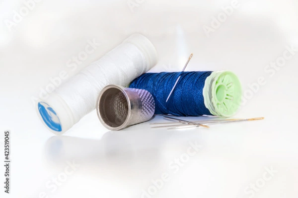 Fototapeta White and blue thread spools with needles and sewing thimble on a brightness white background