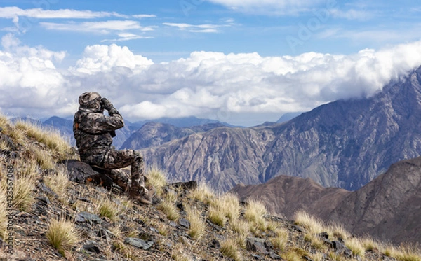 Fototapeta Man in camouflage on the slope is watching through binoculars in the mountains.