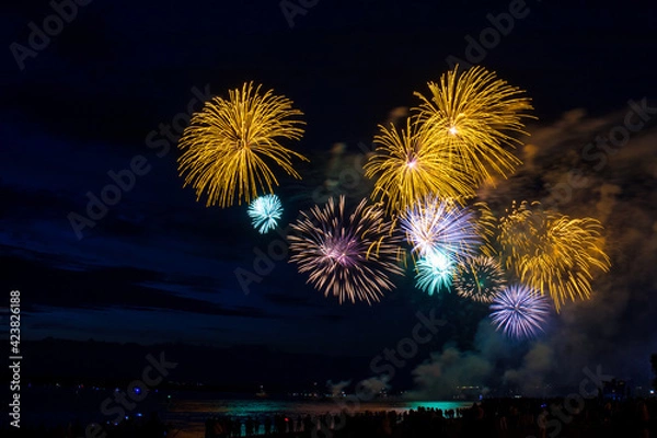 Fototapeta Festive  fireworks launch from a barge on the water, the silhouettes of the audience are visible.