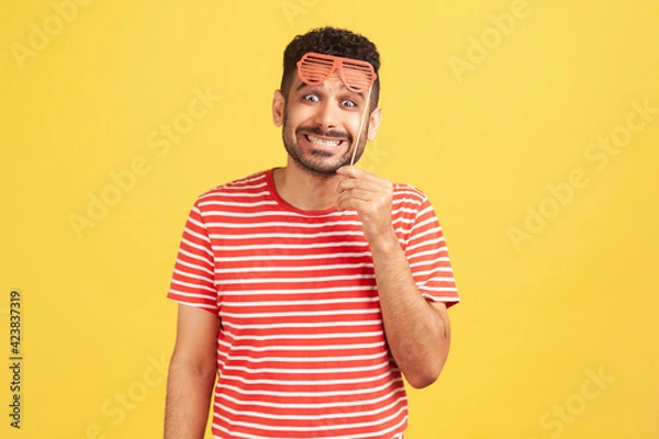 Fototapeta Excited positive man with beard in striped t-shirt holding party mask on stick and looking at camera with toothy smile, having fun. Indoor studio shot isolated on yellow background