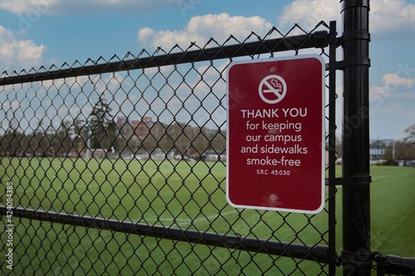 Fototapeta Smoke-free zone sign on chain link fence with university green grass playing field in background