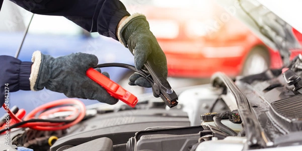 Fototapeta Human hands with jumper cable over the automobile engine in close up.
