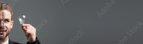 Fototapeta partial view of smiling businessman looking at light bulb isolated on grey, banner