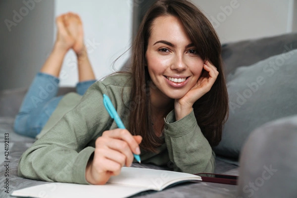 Fototapeta Young smiling woman writing down notes while lying on sofa at home