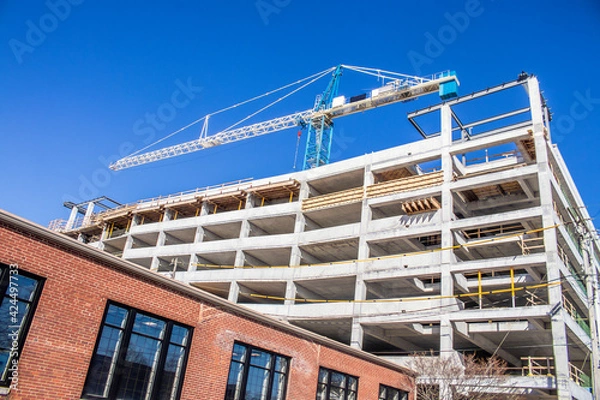 Fototapeta Parking garage under construction - slanted view of concrete bones of building with giant crane on top with lower pre-existing brick building in foreground