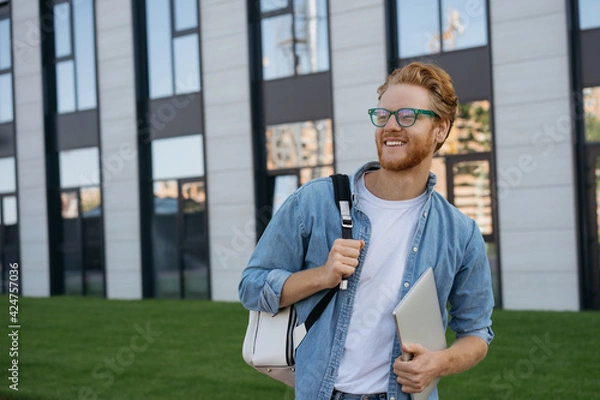 Fototapeta Portrait of happy confident man wearing eyeglasses looking away, copy space. Smiling university student with backpack and laptop computer walking in university campus, education concept 