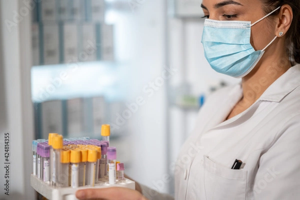 Fototapeta Portrait of smiling woman laborant eith face mask holding test tubes in laboratory.