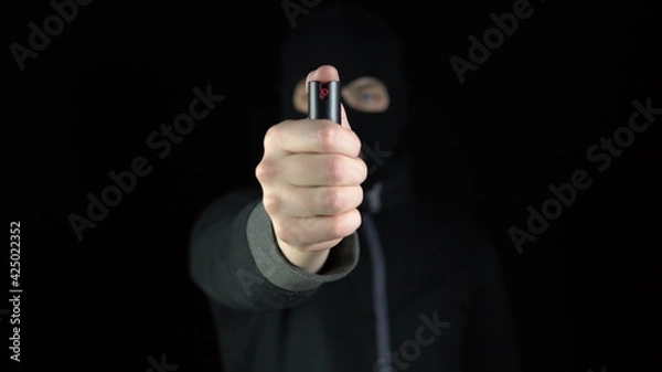 Fototapeta A man in a balaclava mask stands with a pepper spray. A bandit holds out a pepper spray to the camera. On a black background.