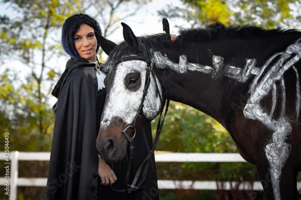 Fototapeta Portrait of a beautiful girl in clothes in a medieval style, next to there is a horse painted with white paint with a painted skeleton