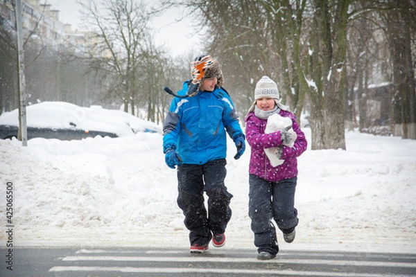 Fototapeta In winter, on the street in snowy weather, a boy and a girl cross the road along a pedestrian crossing.