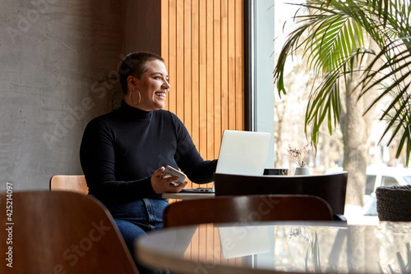 Fototapeta A young girl with short hair and a nose piercing is sitting in a cafe and working on a laptop. The concept of freelancing and remote work or training.