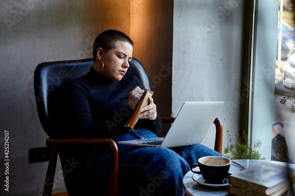 Fototapeta A young girl with glasses, with short hair is sitting in a cafe and working on a laptop. The girl looks at her laptop and makes notes in a notebook. The concept of distance learning