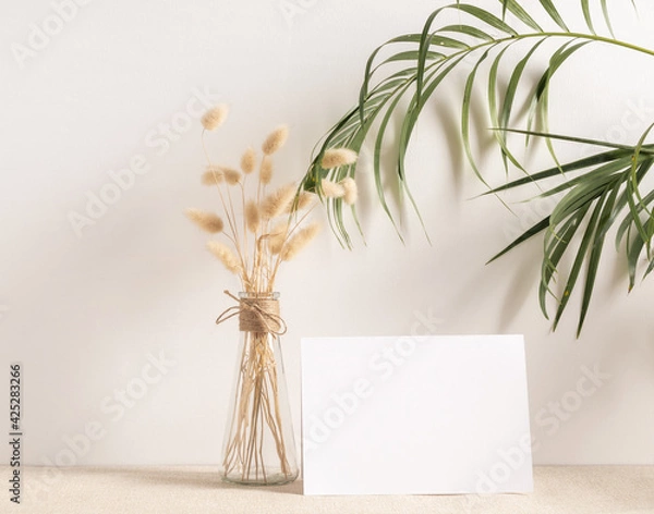 Fototapeta Blank white greeting card mock up.decoration with dried Lagurus ovatus flowers composition in modern glass vase  on  beige table and cement wall background