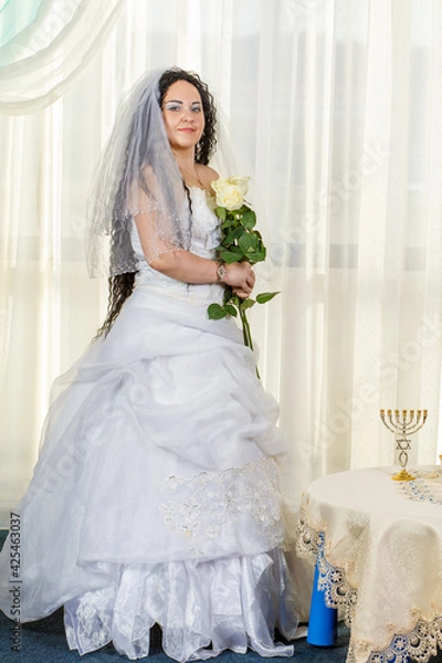 Fototapeta A Jewish bride stands in a synagogue at a table with flowers before a chuppa ceremony during a pandemic, wearing a medical mask and a bouquet of flowers