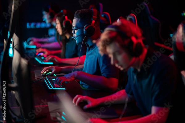 Fototapeta A multi-racial team of esports athletes conducts a training session before an online shooter tournament. Neon light.