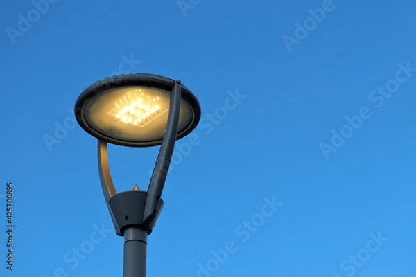 Obraz Led lamp glowing on blue sky background. Electric lighting, energy-saving street lantern