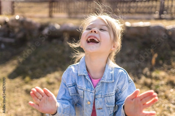 Fototapeta A seven year old girl laughs and rejoices in the spring on the street in a denim jacket.