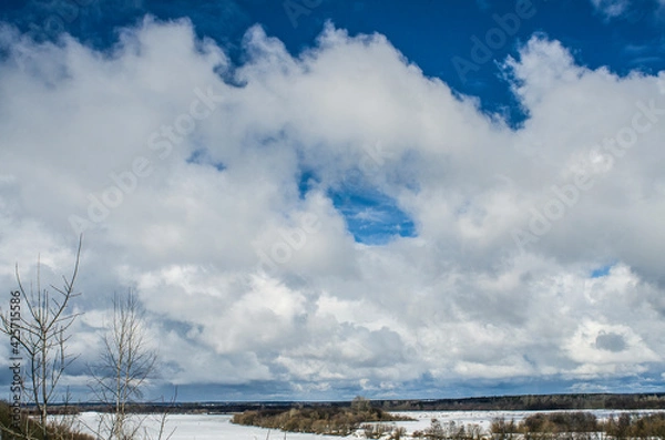 Obraz Winter landscape with a high bank of the river covered with ice, cumulus clouds in the sky,.deep snow cover with footprints, a clear horizon line.