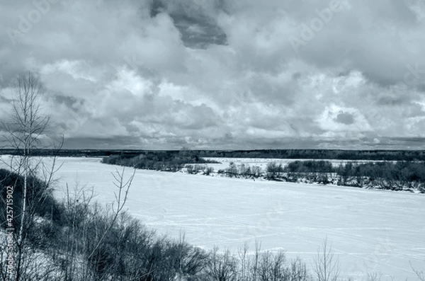 Obraz Winter landscape with a high bank of the river covered with ice, cumulus clouds in the sky,.deep snow cover with footprints, a clear horizon line, monochrome, blue tone.
