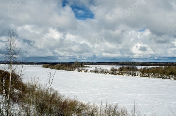Obraz Winter landscape with a high bank of the river covered with ice, cumulus clouds in the sky,.deep snow cover with footprints, a clear horizon line.