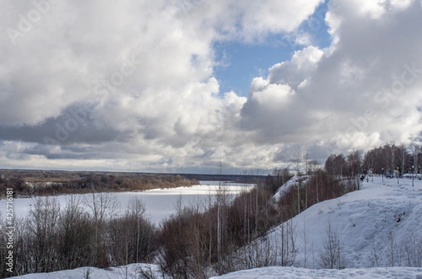 Obraz Winter landscape with a high bank of the river covered with ice, cumulus clouds in the sky,.deep snow cover with footprints, a clear horizon line.