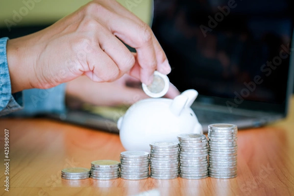 Obraz Business men put the coins into the white piggy bank and the coins stacks were lined up in front. The idea of ​​saving money to be used in the future. Financial and business concept