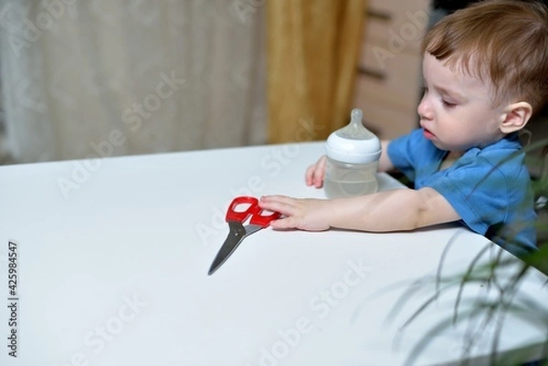 Fototapeta Danger in the home, sharp objects are dangerous for young children. little boy picks up scissors from the table and can hurt himself