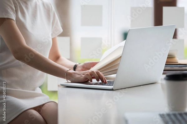 Fototapeta Cropped photo hand of woman writing making list taking notes in notepad working or learning online with laptop at home