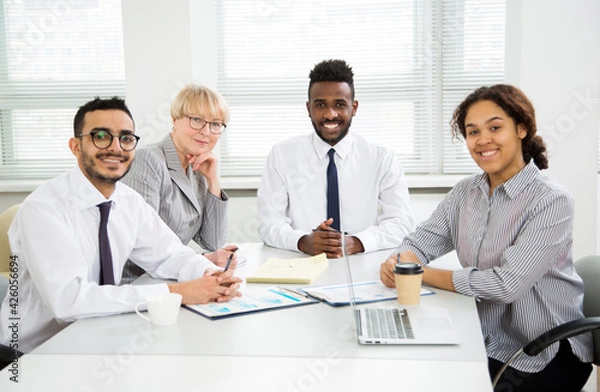 Fototapeta Multy-ethnic group of business people working while sitting at the office desk and smiling at camera