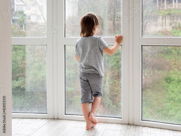 Fototapeta Little boy looks outdoors through wet panoramic window with raindrops. Rainy summer weather. Kid stays alone at home. Loneliness and sad mood without children's company.