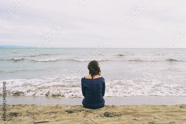 Fototapeta girl sitting on the sand on the shore of the beach looking at the horizon of the sea
