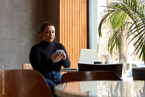 Fototapeta A young girl with short hair and a nose piercing is sitting in a cafe and working on a laptop. The concept of freelancing and remote work or training.