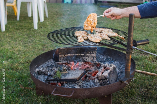 Fototapeta Fire ring and poultry meat on grill and human hand with fork. Grilling season and summer.