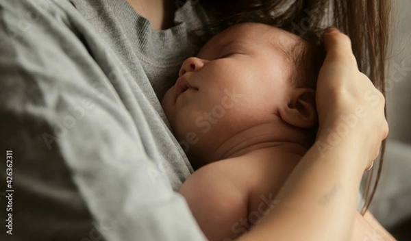 Obraz Authentic young mother holds newborn baby in her arms on to the chest, strokes his hair while he sleeps. Tenderness and care for the child. Maternity and motherhood concept.